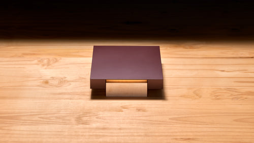 Vora Aegis on a pale wood floor under a warm light: a burgundy aluminum slab resting on a solid wood block